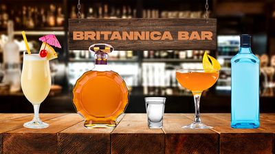 A piña colada, a bottle of brown liquor, a full shot glass, a cocktail in a coupe glass, and a bottle of gin sit on a bar. A sign labeled Britannica Bar hangs above them.