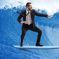 Man in a black suit and tie surfing on a blue surfboard in front of a large ocean wave.